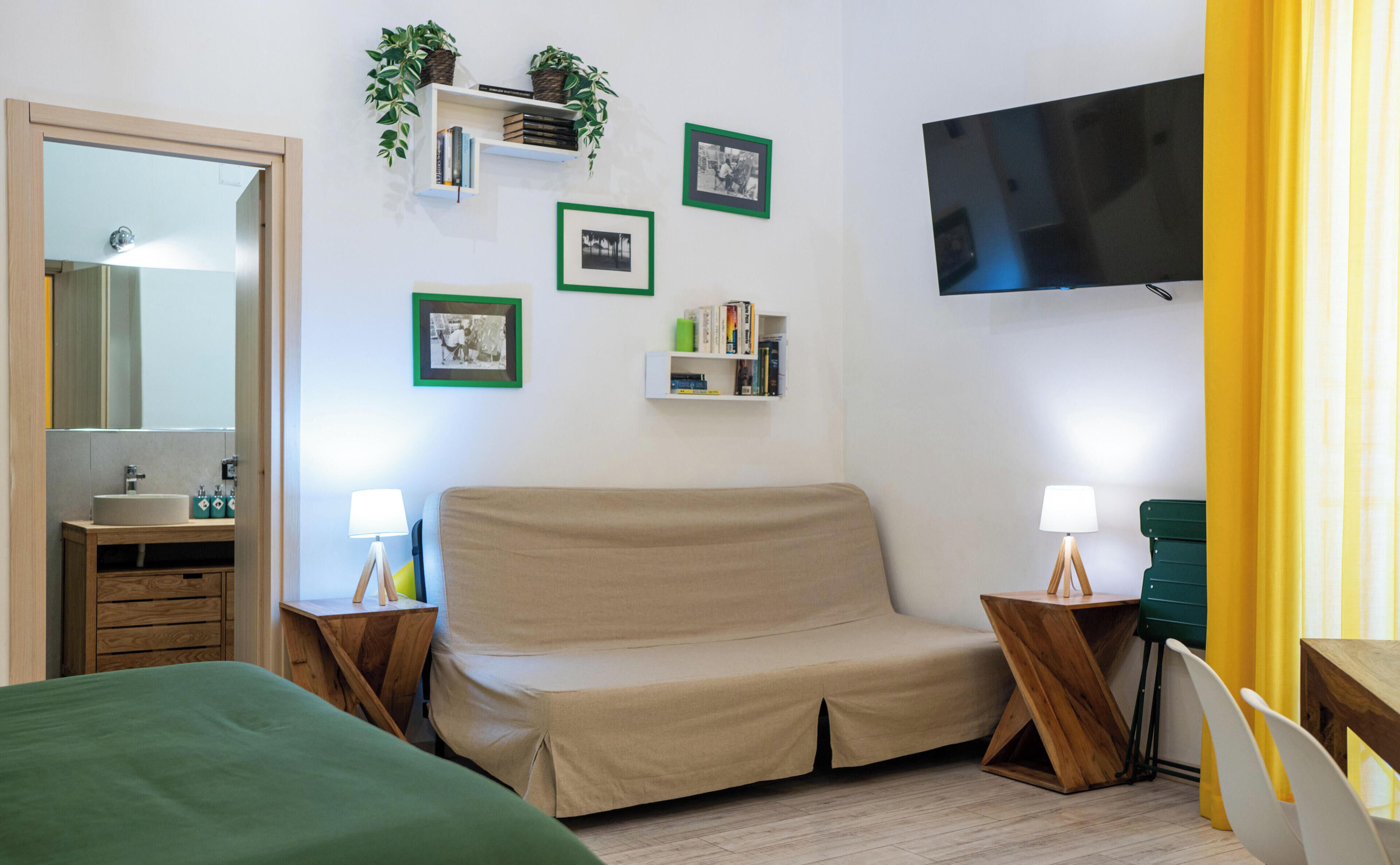A bedroom with a bed, a sofa, a TV, a mirror, and a bedside table with a lamp.