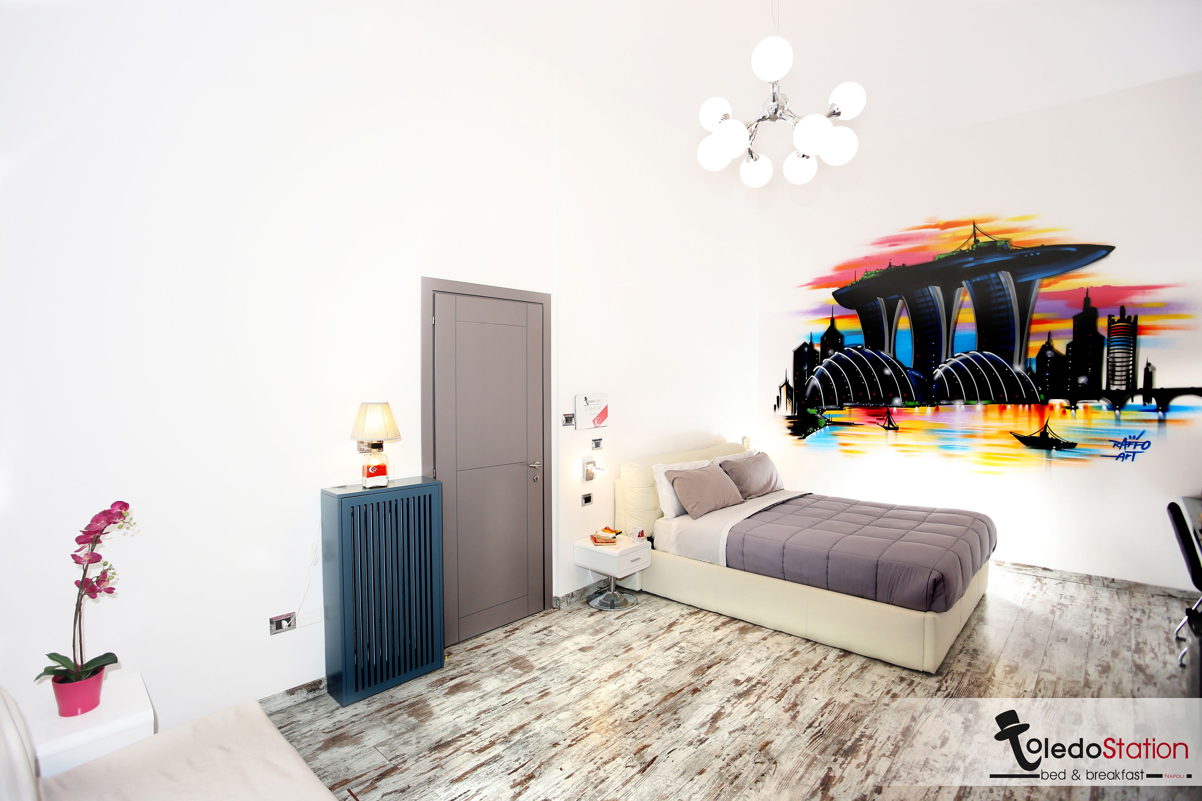 A modern bedroom with a bed, bedside table, lamp, and a cityscape mural on the wall.
