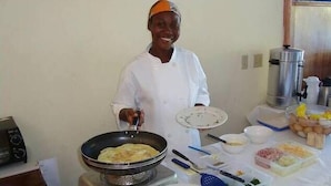 A chef in a white uniform cooking on a portable stove, with various food items and kitchen utensils on the table. - Cap Lamandou Hotel (Jacmel)