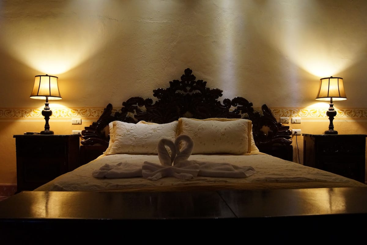A neatly made bed with a decorative headboard, two bedside lamps, and a pair of towels arranged in a heart shape on the bed.