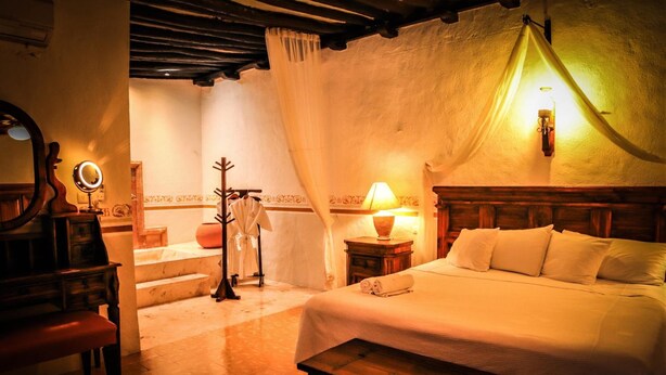 Un dormitorio con cama, mesita de noche, lámpara, espejo, banco y perchero.