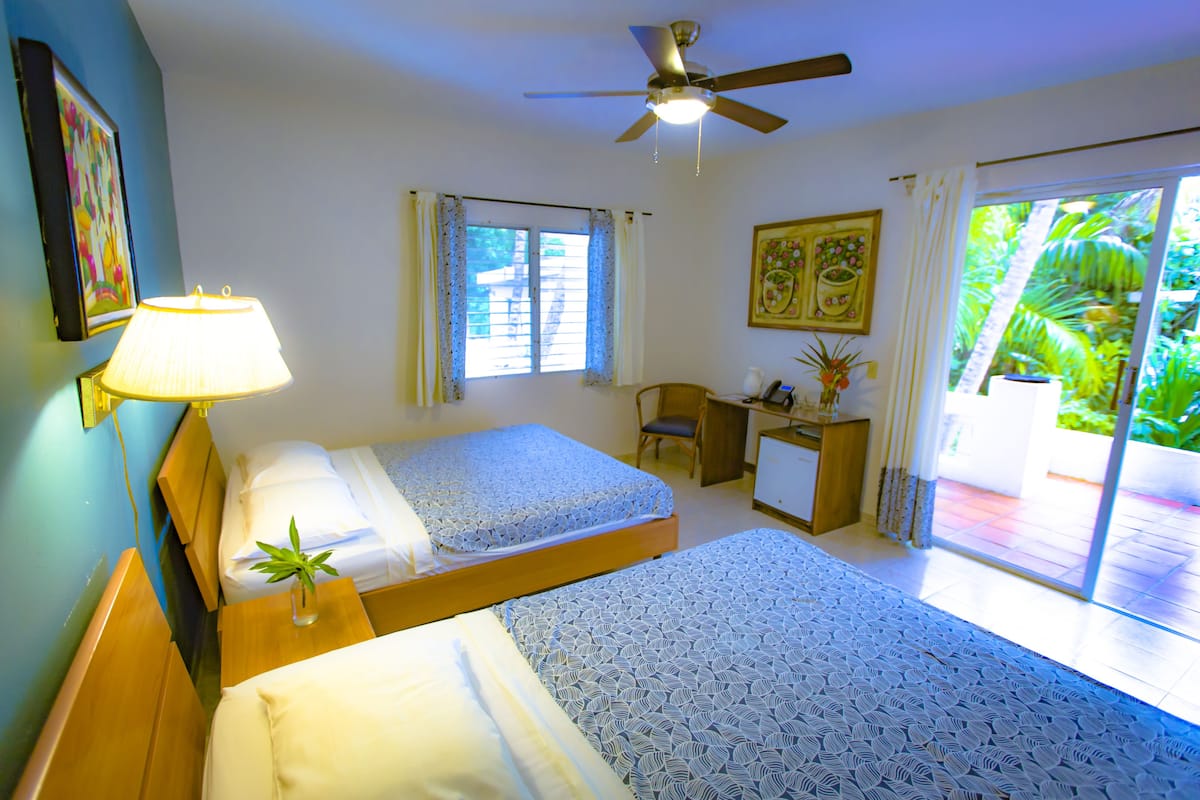 A hotel room with two beds, a desk, a ceiling fan, and a view of the outdoors.