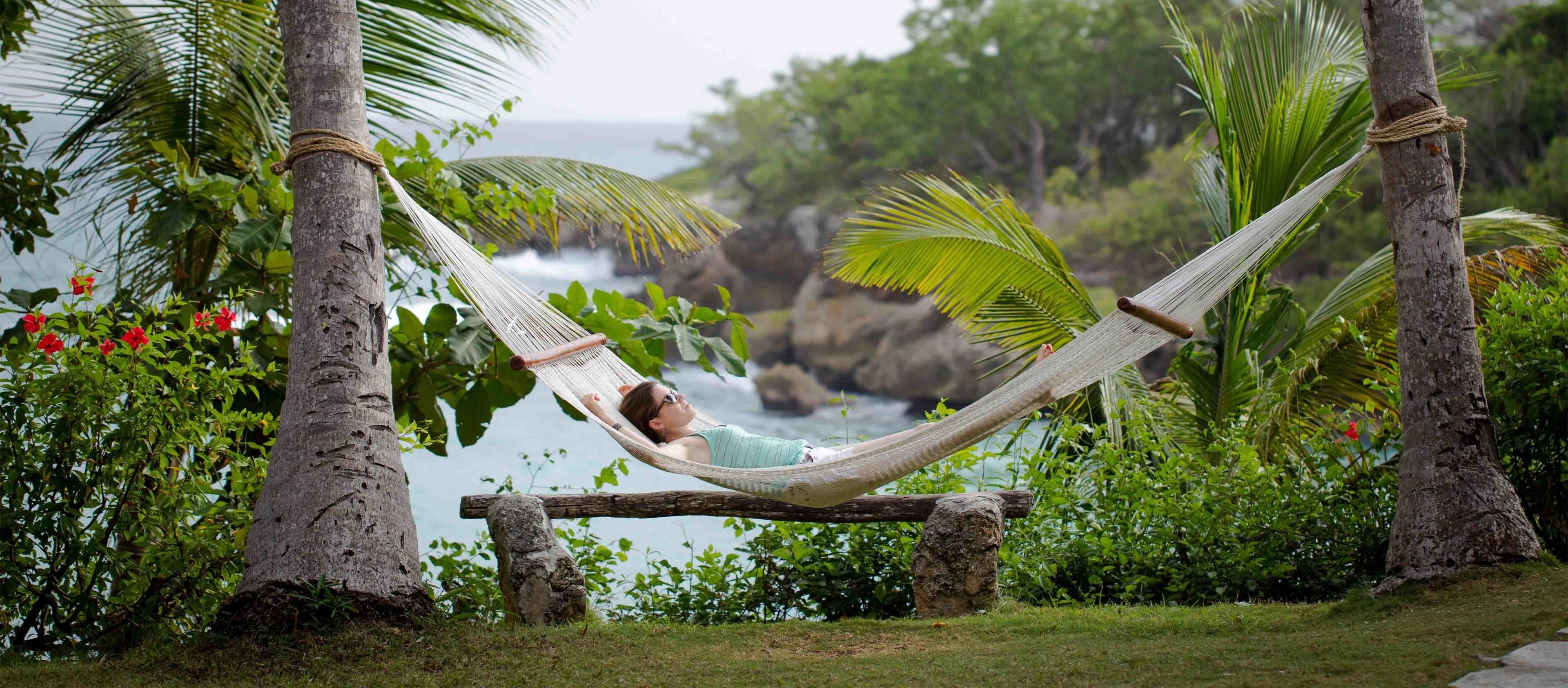 A hammock tied between trees with a person lying in it, overlooking a rocky coastline and lush greenery.