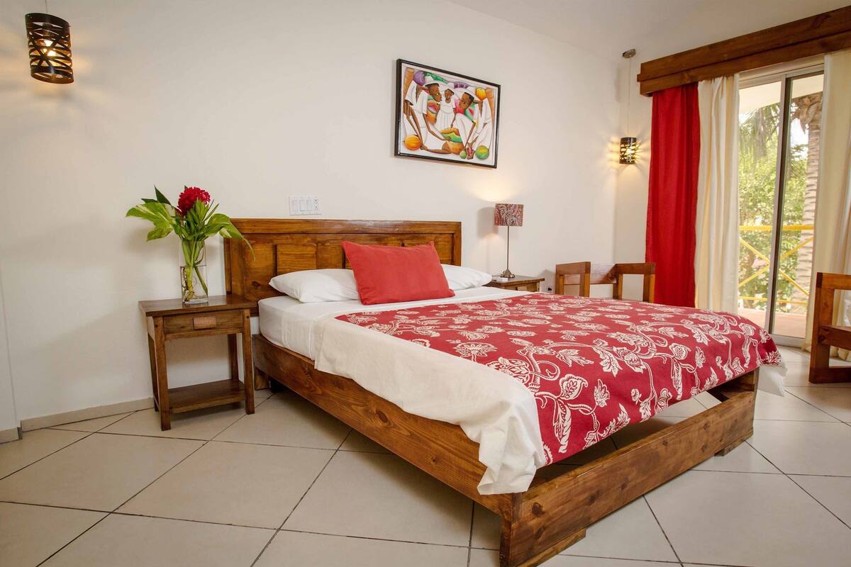 A wooden bed with a red and white bedspread, a nightstand with a vase of flowers, a wall-mounted painting, and a window with red curtains.