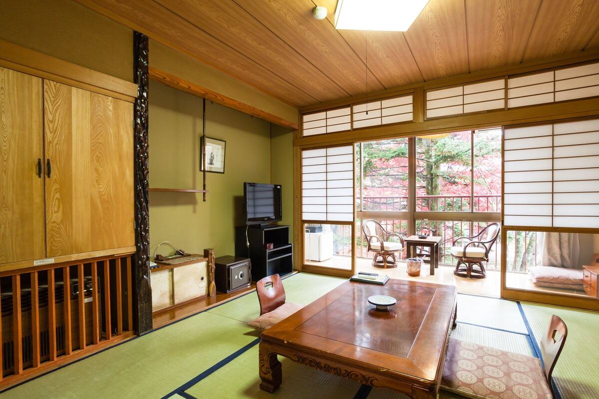 A traditional Japanese room with tatami flooring, a wooden table, tatami mats, a television, and a view of cherry blossoms.