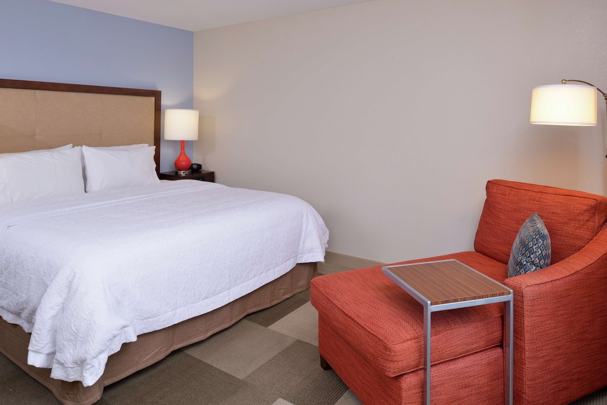 A hotel room with a large bed, a nightstand with a lamp, a red armchair, and a small table.