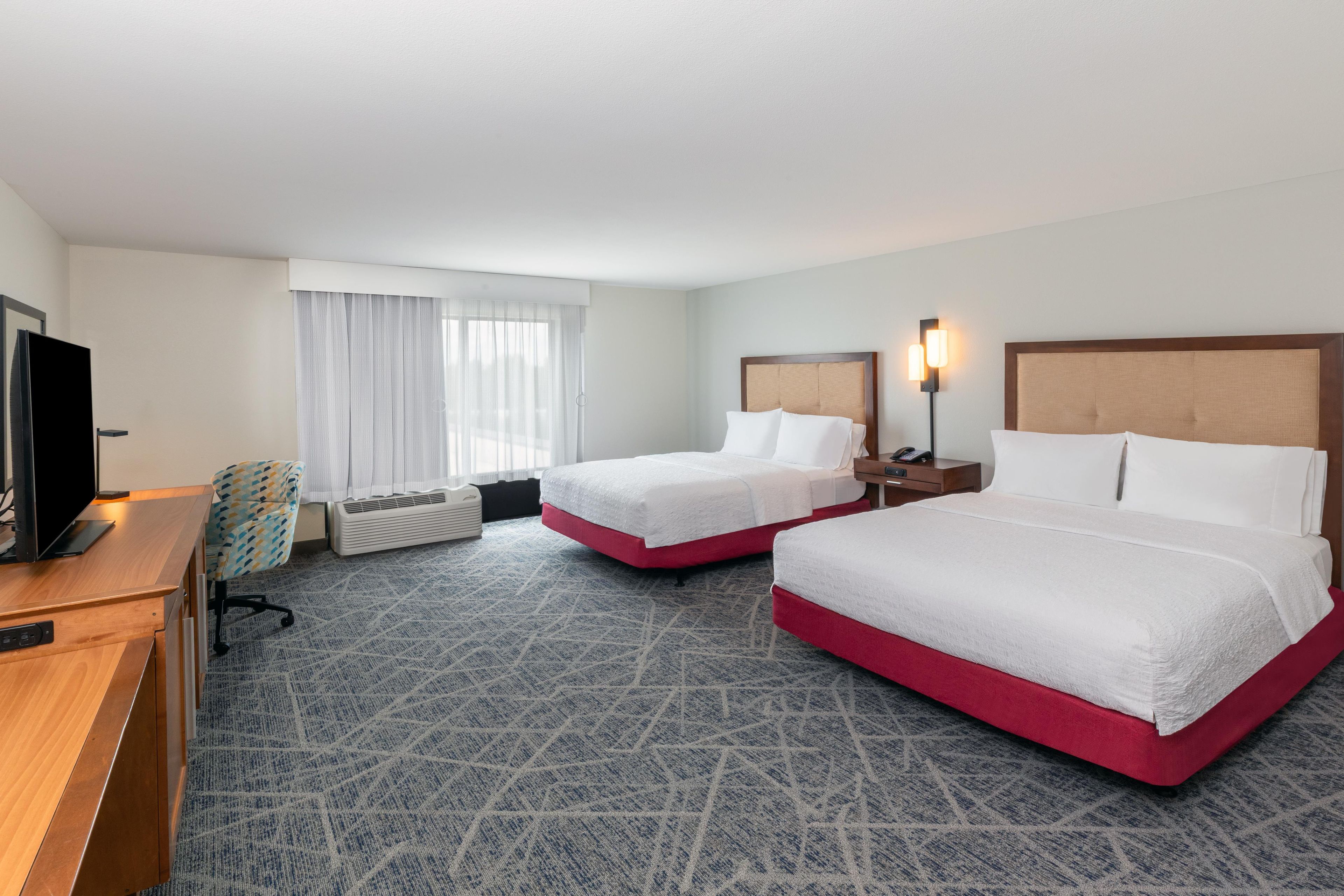 A hotel room with two beds, a desk, a television, and a window with curtains.