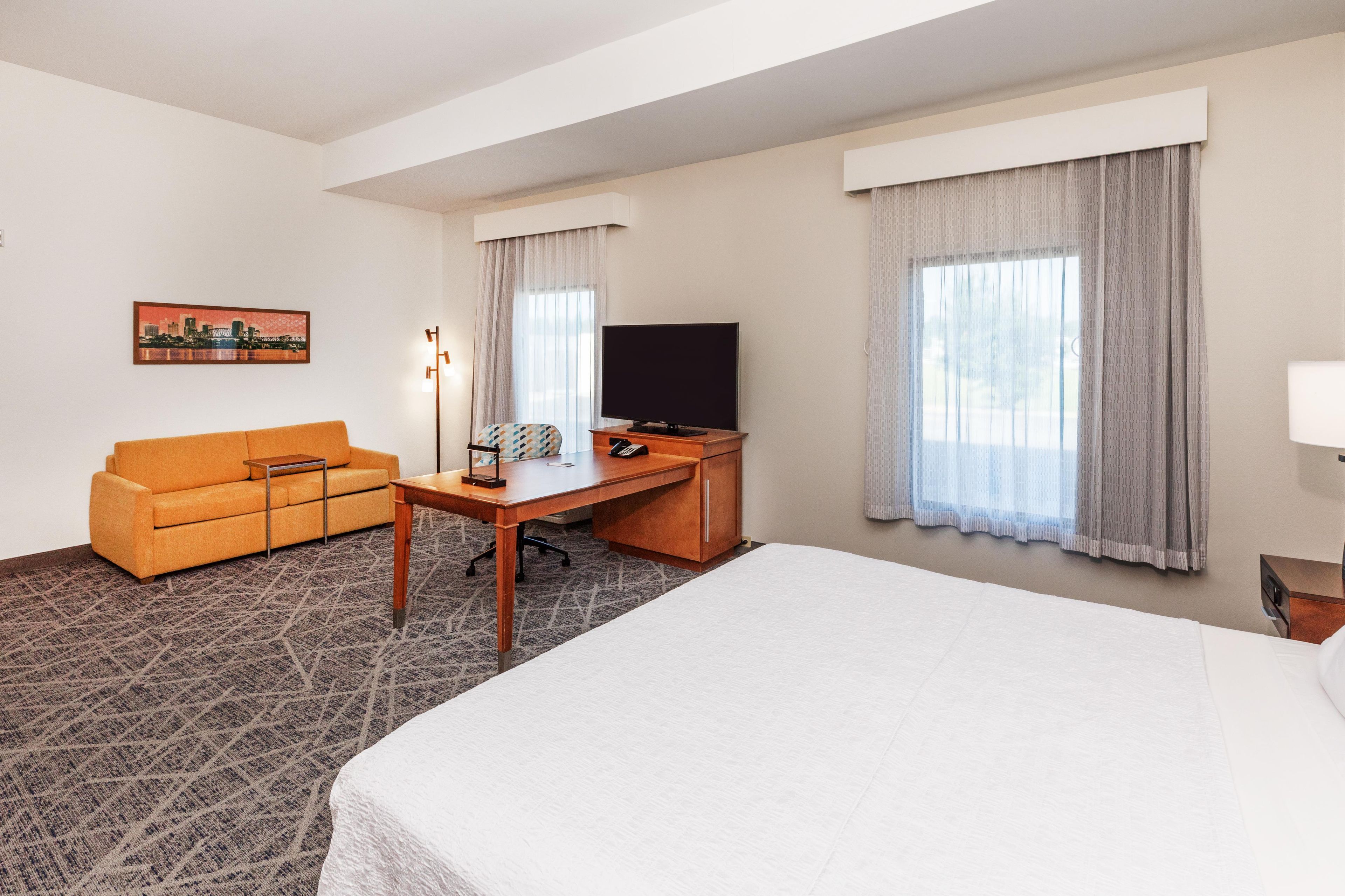 A hotel room with a bed, a sofa, a desk with a TV, and a cityscape painting.