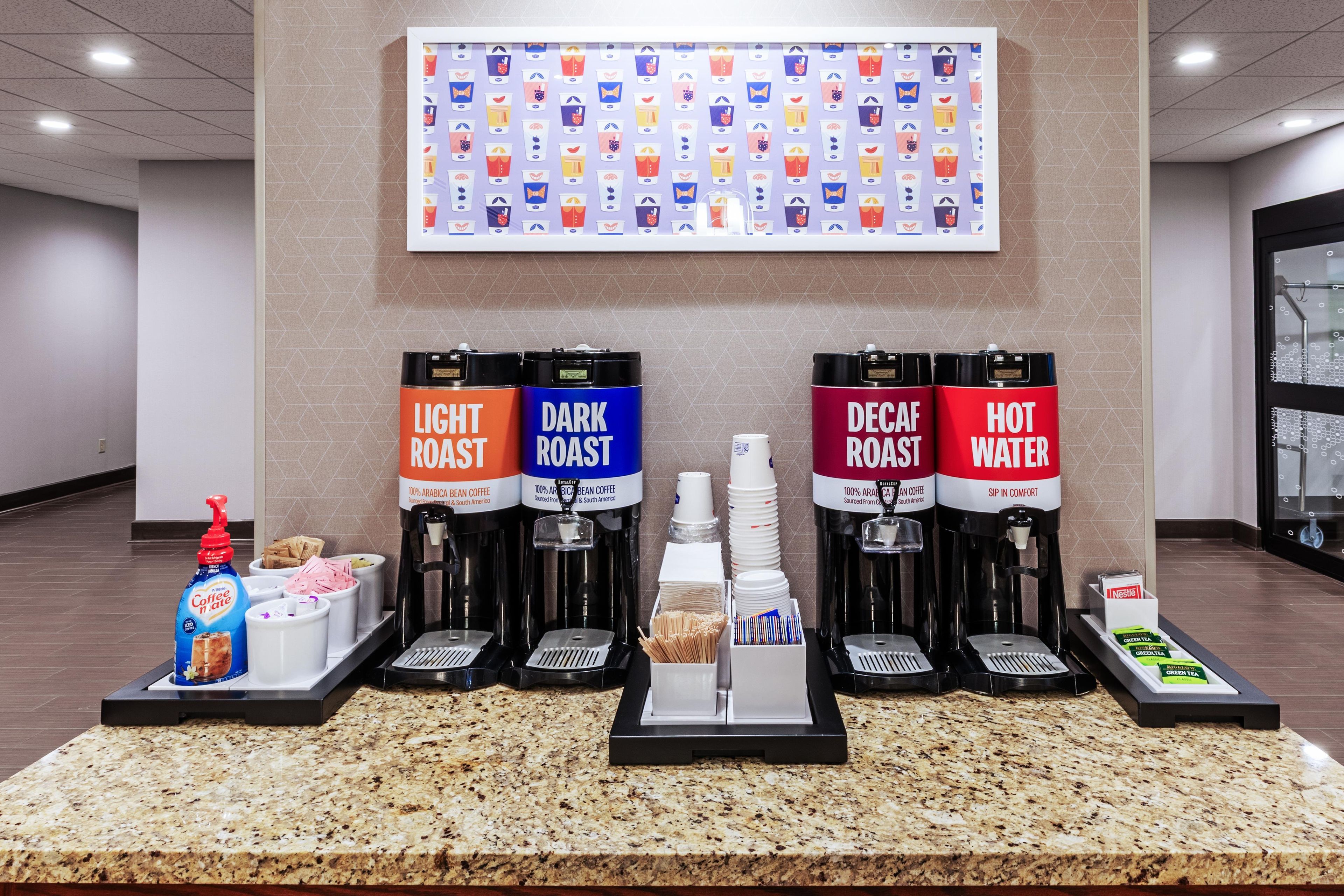 Una estación de café con cuatro dispensadores de café etiquetados como 'Light Roast', 'Dark Roast', 'Decaf Roast' y 'Hot Water'. También hay cápsulas de café y una botella de crema.