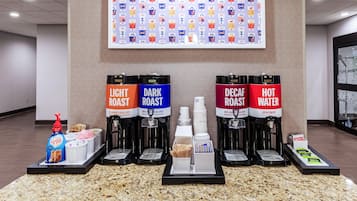 Una estación de café con cuatro dispensadores de café etiquetados como 'Light Roast', 'Dark Roast', 'Decaf Roast' y 'Hot Water'. También hay cápsulas de café y una botella de crema.