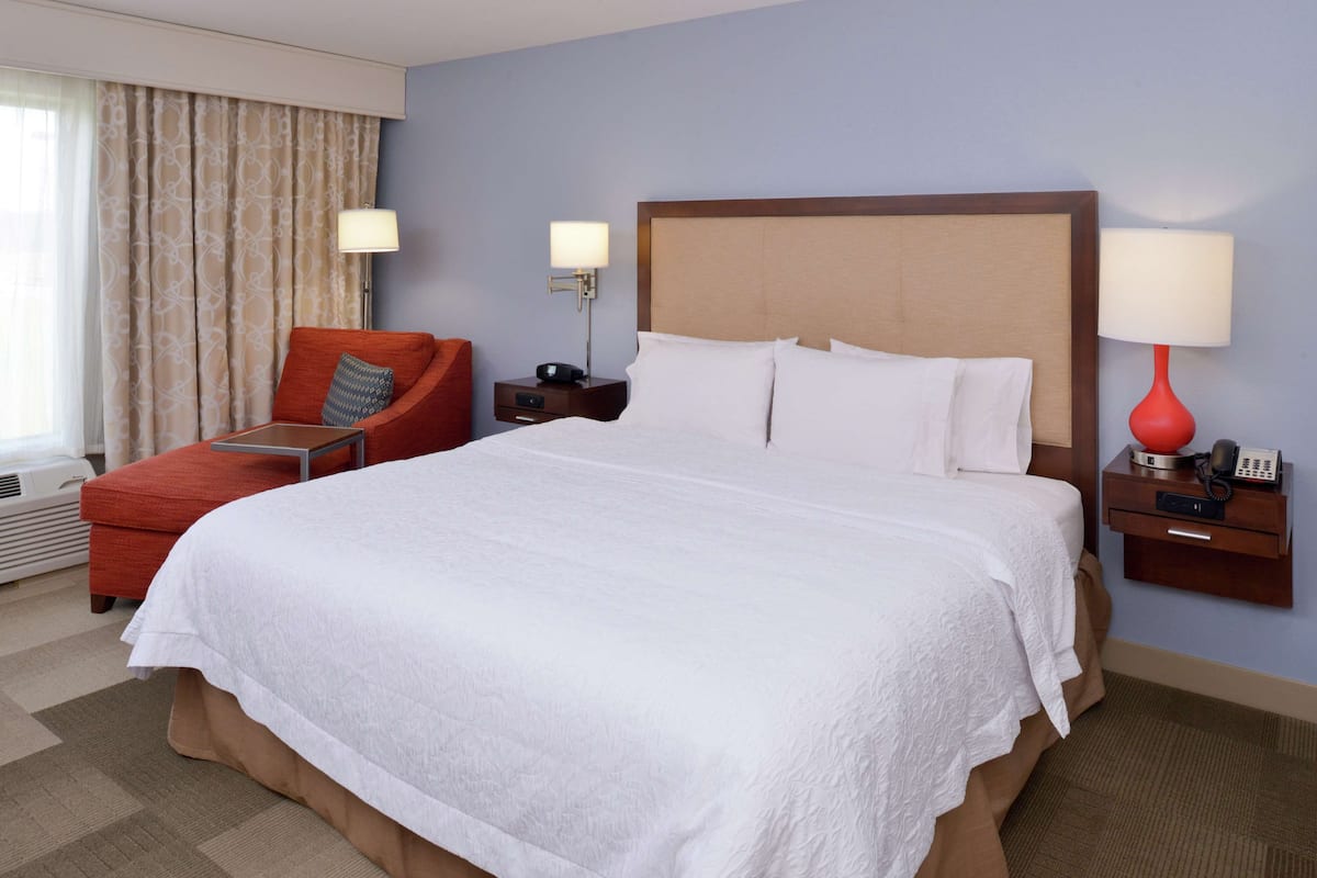 A hotel room with a large bed, a red armchair, two bedside tables with lamps, and a window with curtains.