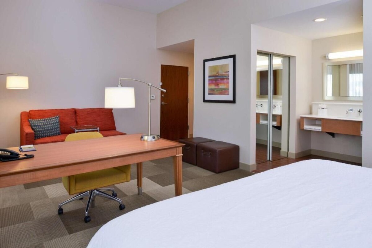 Une chambre d’hôtel avec un lit, un bureau avec une lampe, un canapé avec un coussin, une petite table, une valise, une salle de bain avec un miroir et un tableau encadré au mur.