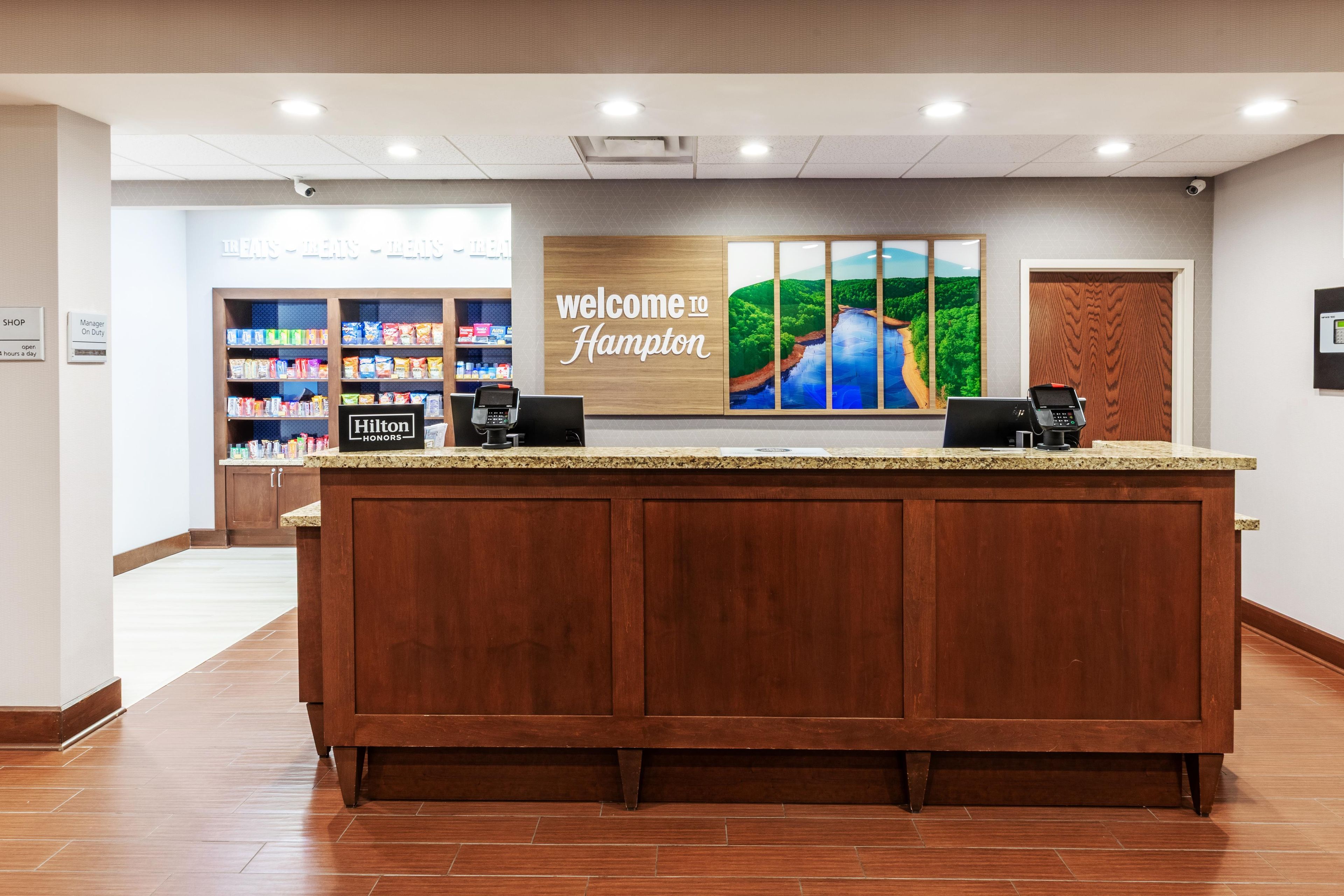 A hotel reception desk with a 'Welcome to Hampton' sign, a 'Hilton Honors' logo, and a view of the hotel's interior including a snack bar.