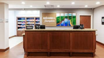 A hotel reception desk with a 'Welcome to Hampton' sign, a 'Hilton Honors' logo, and a view of the hotel's interior including a snack bar.