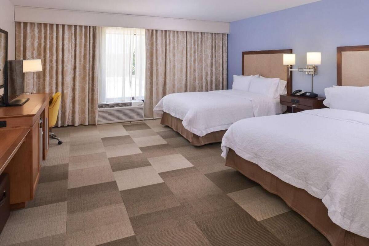 Habitación de hotel con dos camas, un escritorio, una silla, un televisor, una lámpara y una ventana con cortinas.