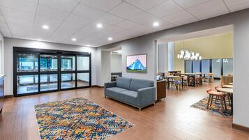 A spacious lobby with a grey sofa, patterned rugs, a large window, and a dining area with tables and chairs.