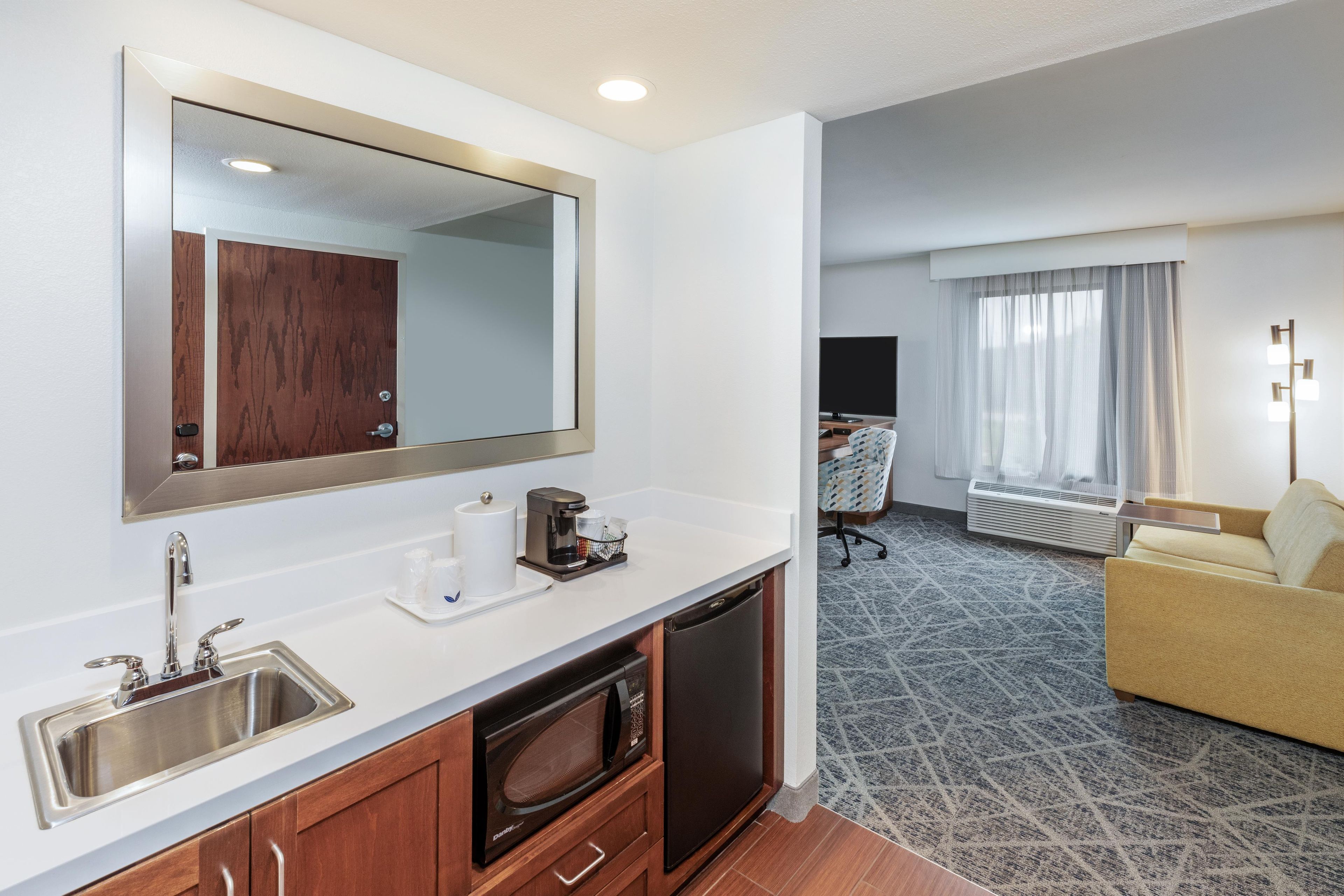 Une chambre d’hôtel équipée d’une kitchenette, d’un grand miroir, d’un lavabo, d’un four à micro-ondes, d’une machine à café et d’un coin salon avec un canapé et une télévision.