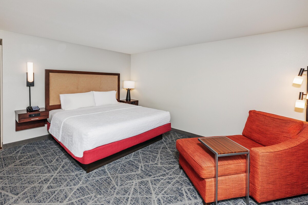A hotel room with a bed, bedside tables, a lamp, a red chair, and a small table.