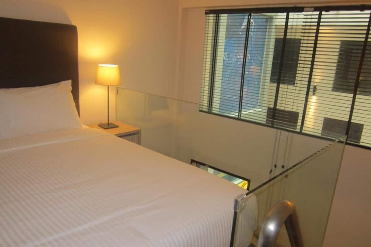 Habitación de hotel con una cama, una mesita de noche con lámpara, barandilla de cristal y ventana con persianas.