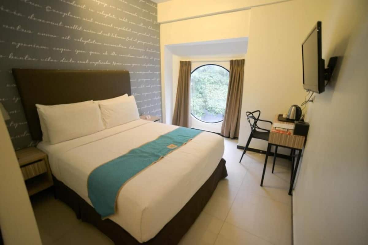Habitación de hotel con cama, escritorio, silla, televisor y ventana con cortinas.