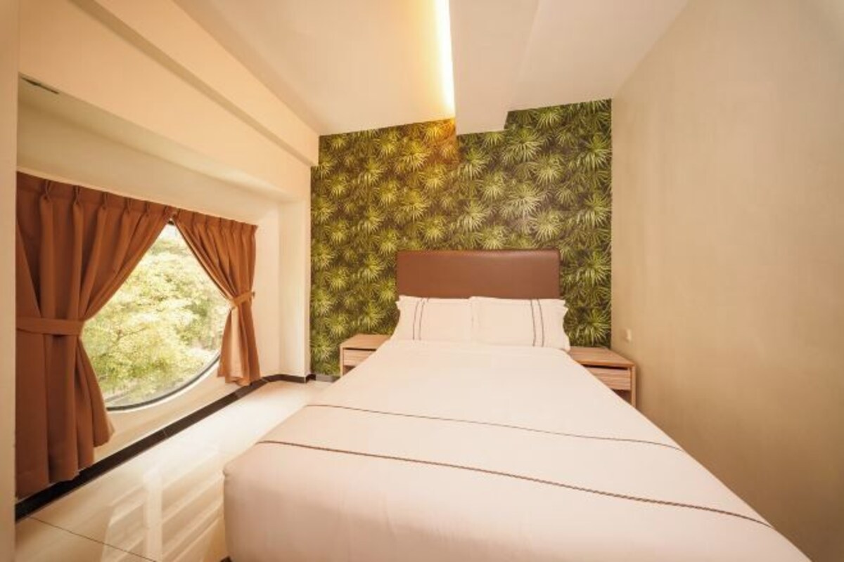 A bedroom with a bed, a nightstand, a window with curtains, and a wall with a leafy pattern.