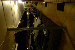 A narrow corridor with a staircase leading upwards, a white vehicle parked on the right, and a metal railing.