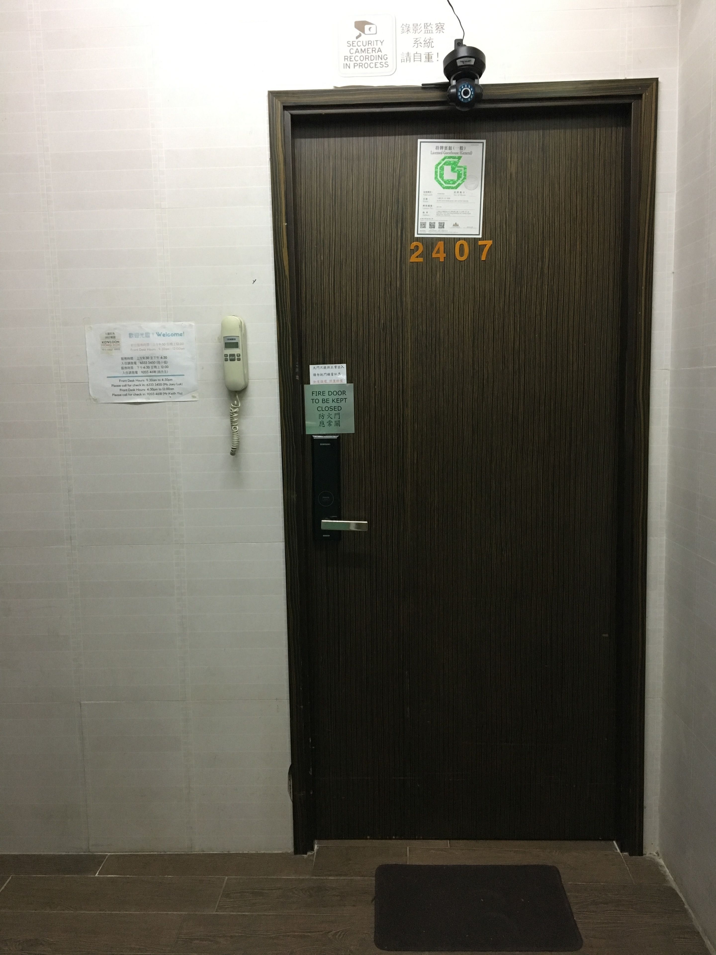Entree van een hotelkamer met een houten deur waarop het cijfer 2407 staat, een beveiligingscamera, een bord met daarop een vluchtdeur en een bord met een telefoonnummer.