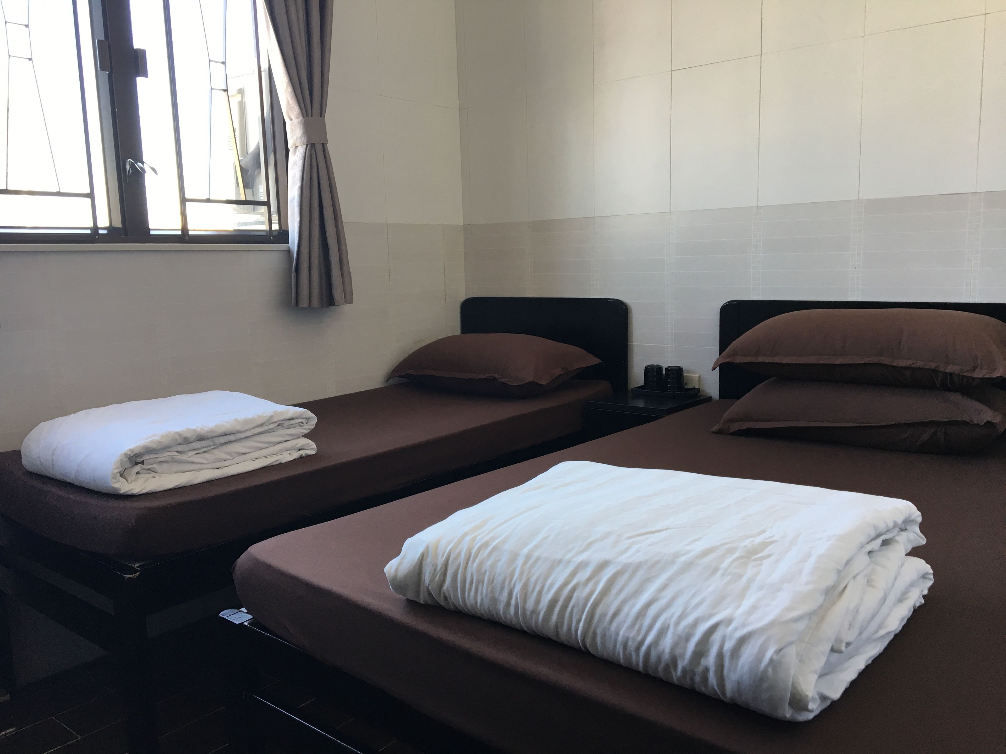 Ein Zimmer mit zwei Einzelbetten, jeweils mit brauner Kopfteil und weißer Decke. Ein Fenster mit Vorhängen, und die Wände sind mit Fliesen versehen.