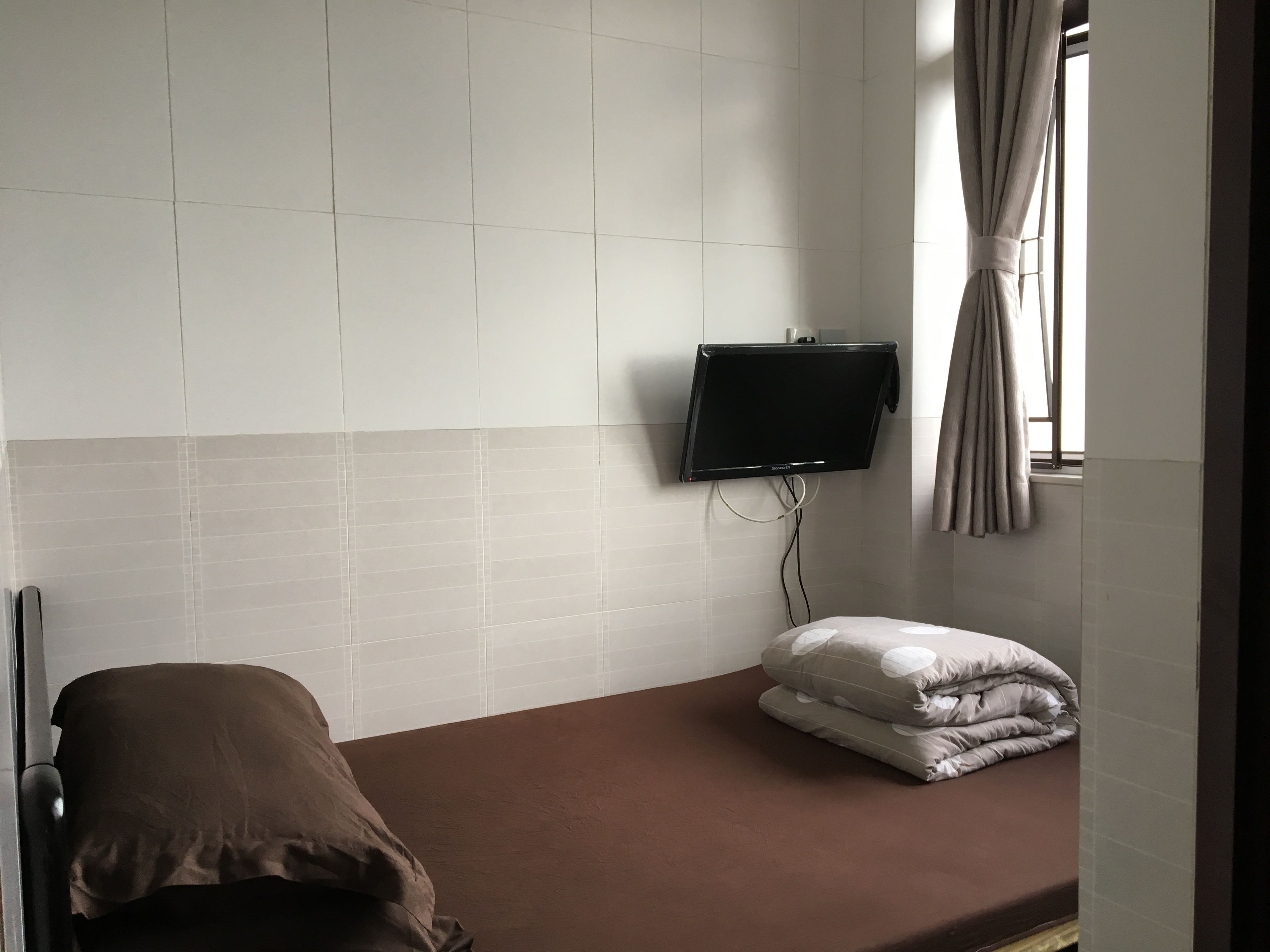 A room with a bed, a television mounted on the wall, and a window with curtains.
