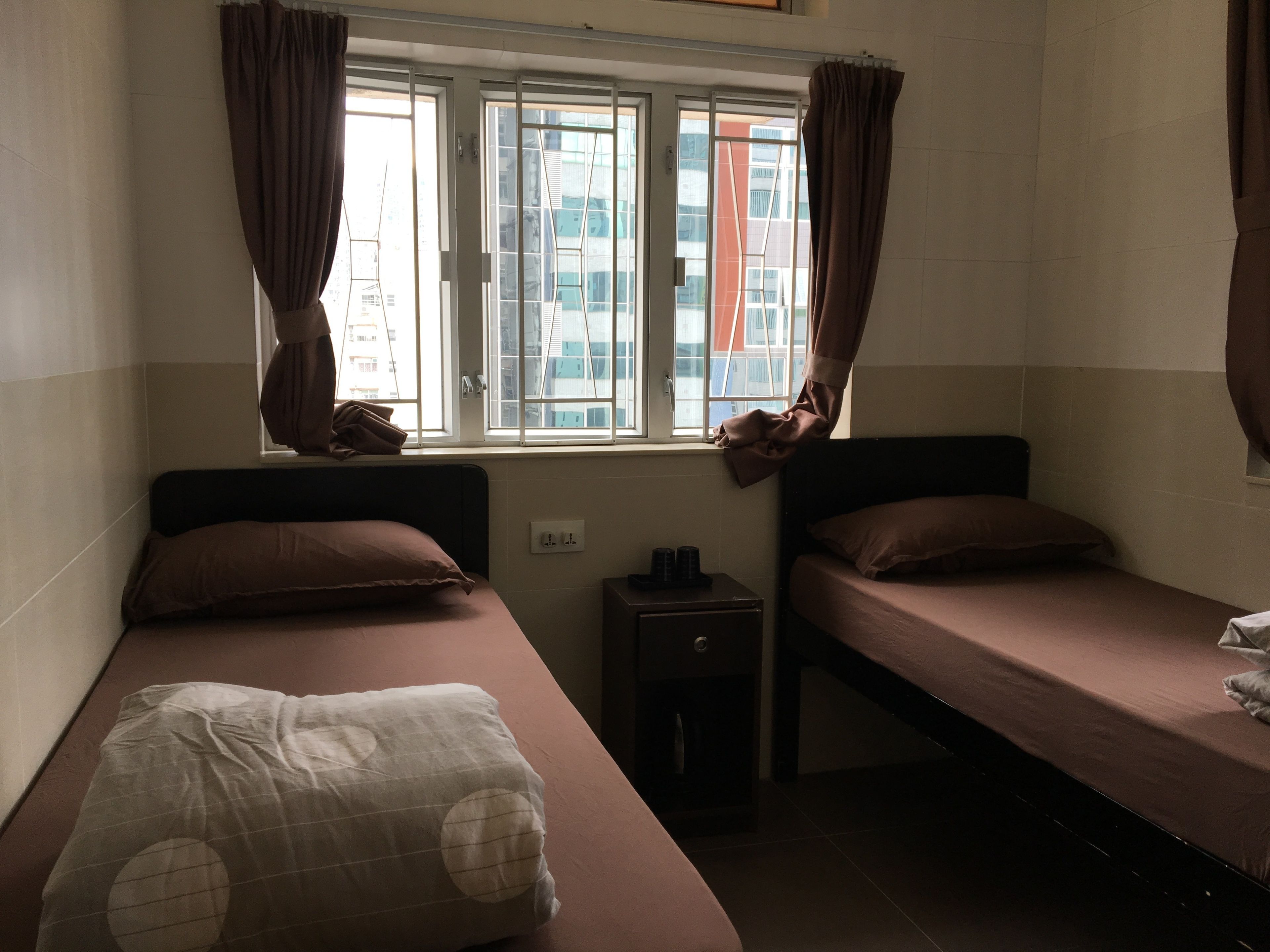 Ein Zimmer mit zwei Einzelbetten, einem Fenster mit Vorhängen, einem kleinen Nachttisch und einer Steckdose.