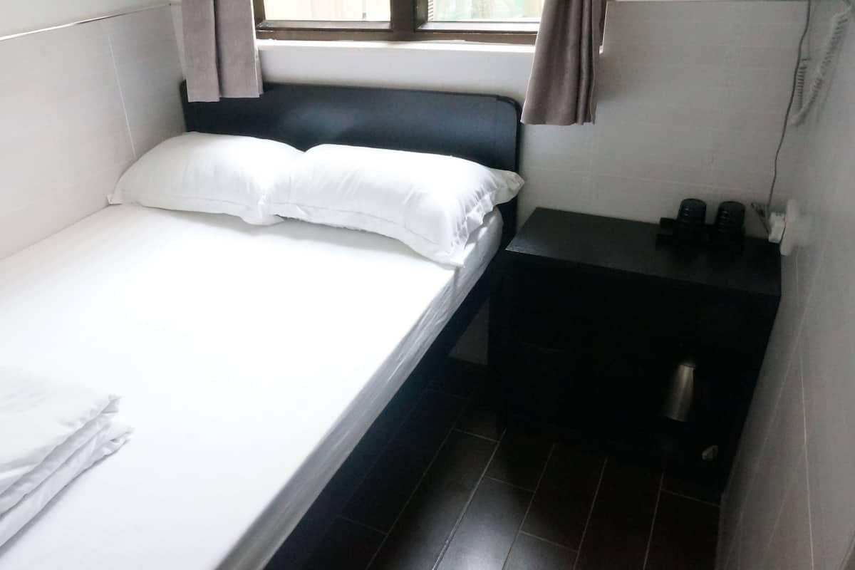 Una cama individual con ropa de cama blanca en una habitación pequeña con cabecera oscura y una ventana con cortinas.
