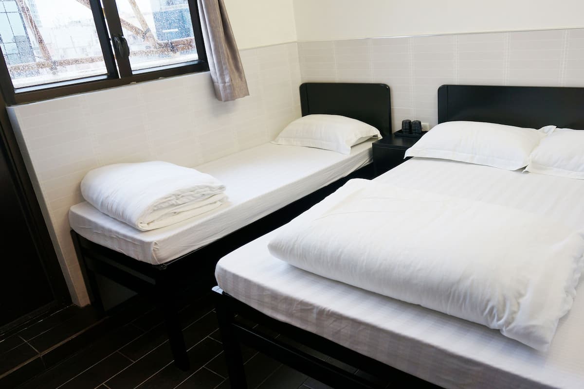 Habitación de hotel con dos camas individuales, una ventana con cortinas, una mesita de noche con lámpara y una pared con azulejos.