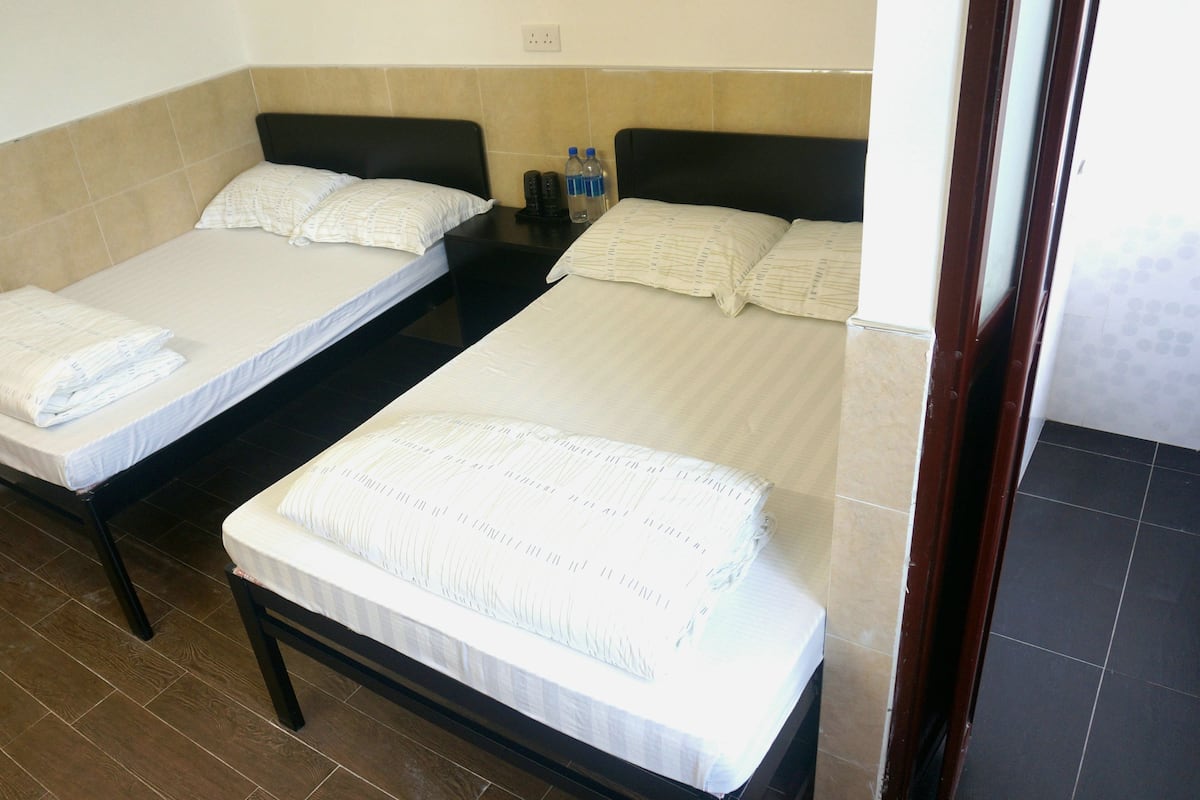 Habitación de hotel con dos camas individuales, cada una con colchón y almohada blancos. Hay dos botellas sobre la mesita de noche entre las camas.