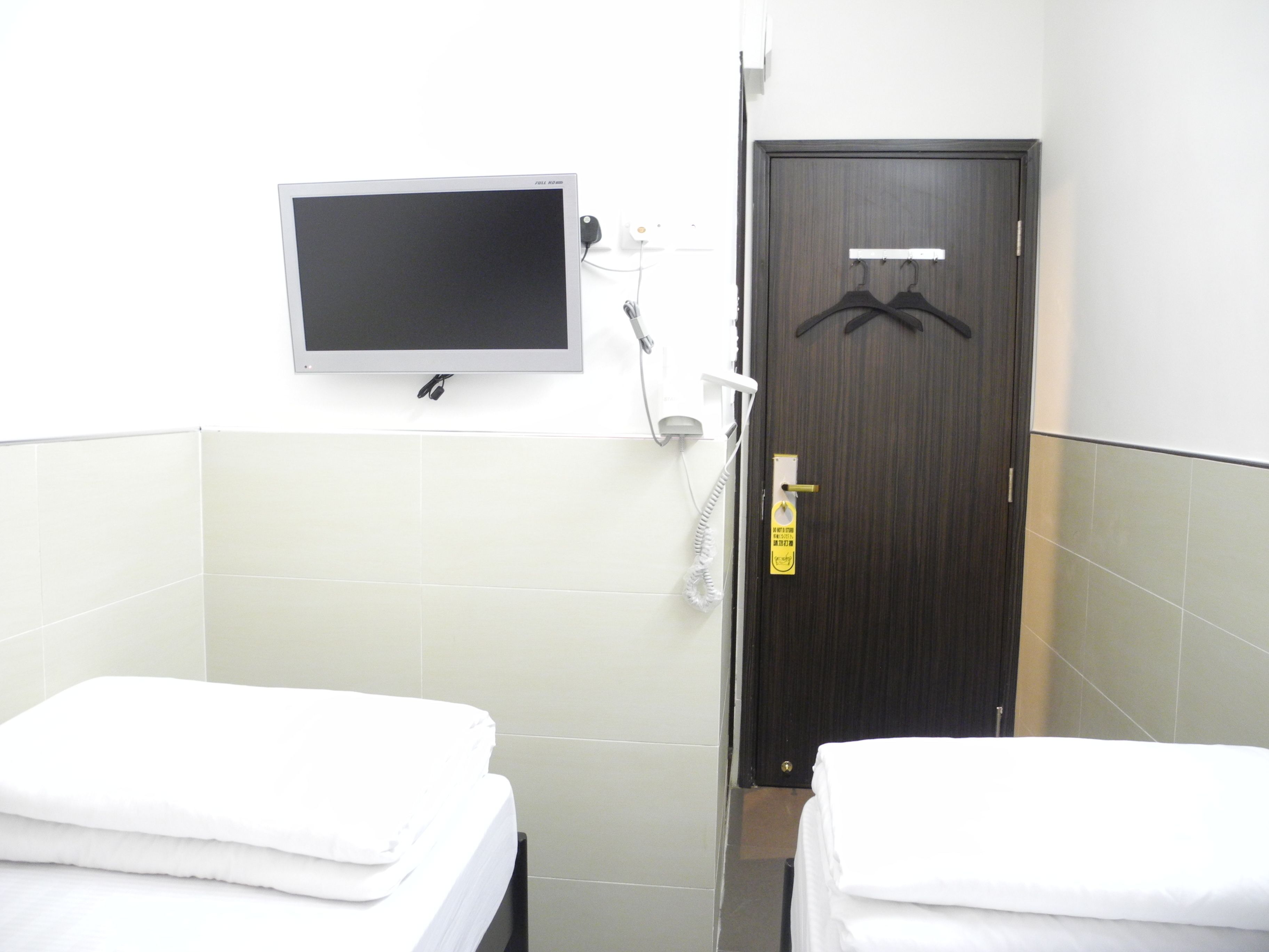 A small, clean room with two single beds, a TV mounted on the wall, and a door with a coat hanging on it.