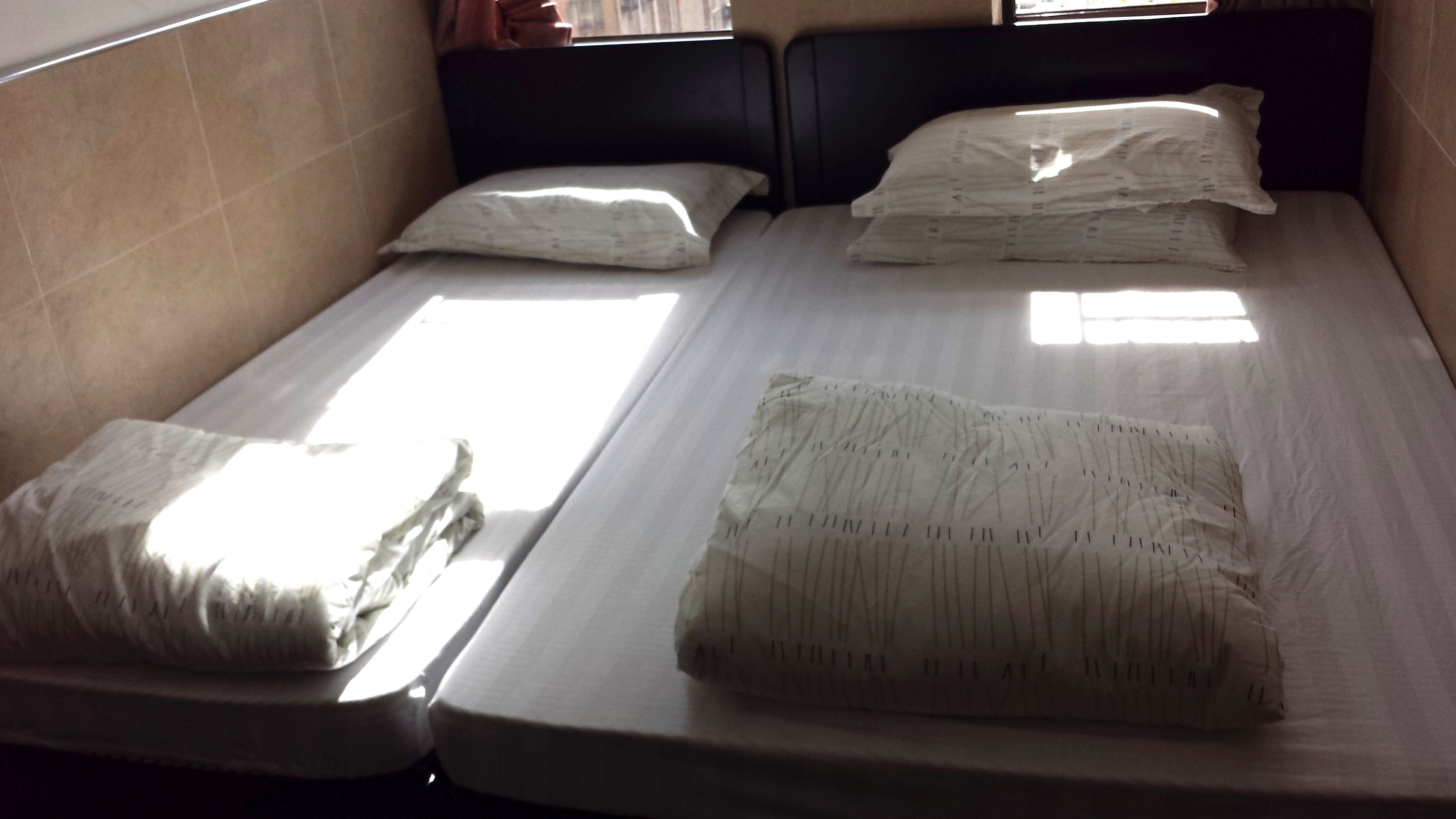 A single bed with a white mattress and two pillows, a window with blinds, and a plain wall.
