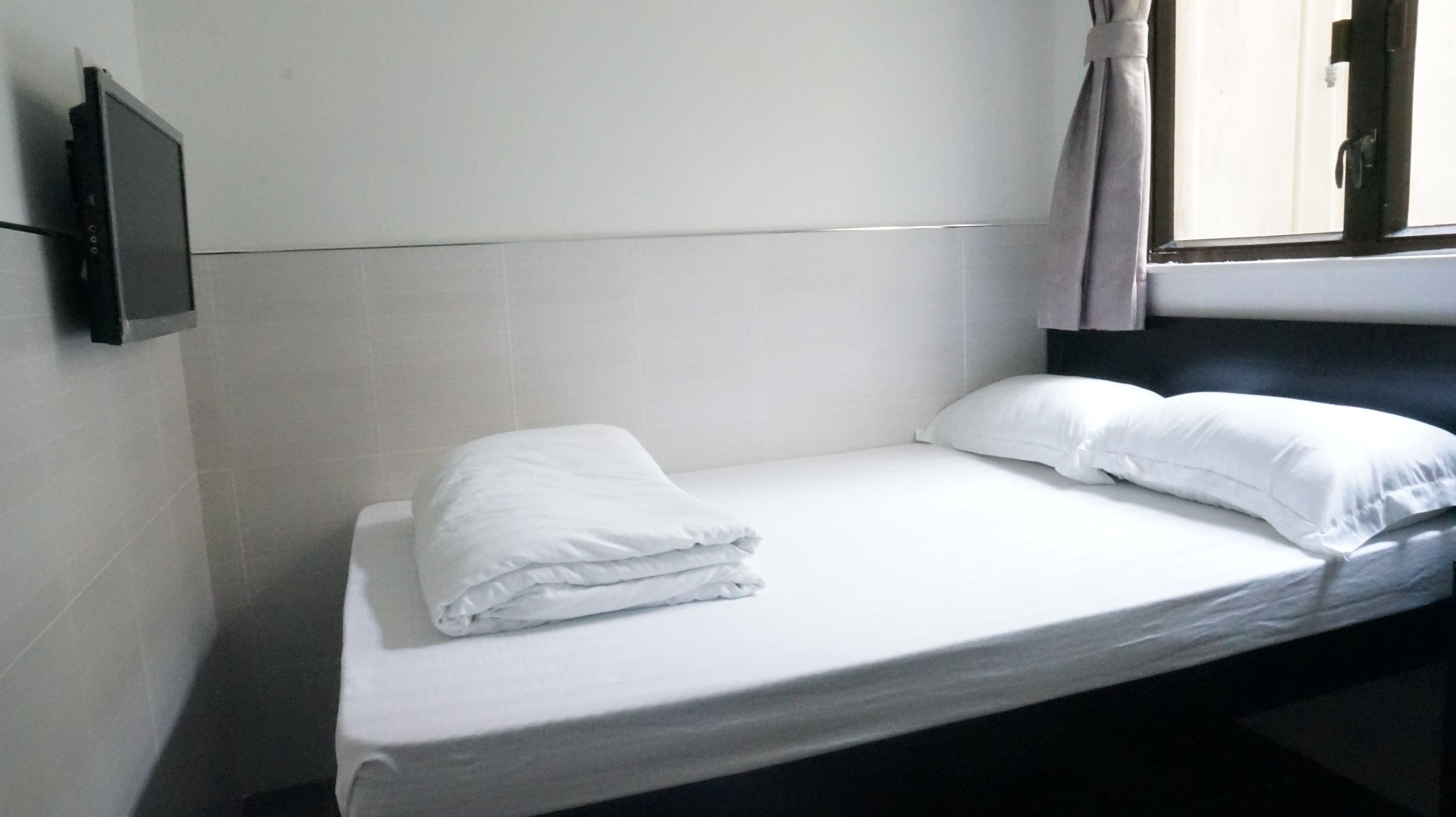A single bed with white linens and pillows, a flat-screen TV mounted on the wall, and a window with curtains.