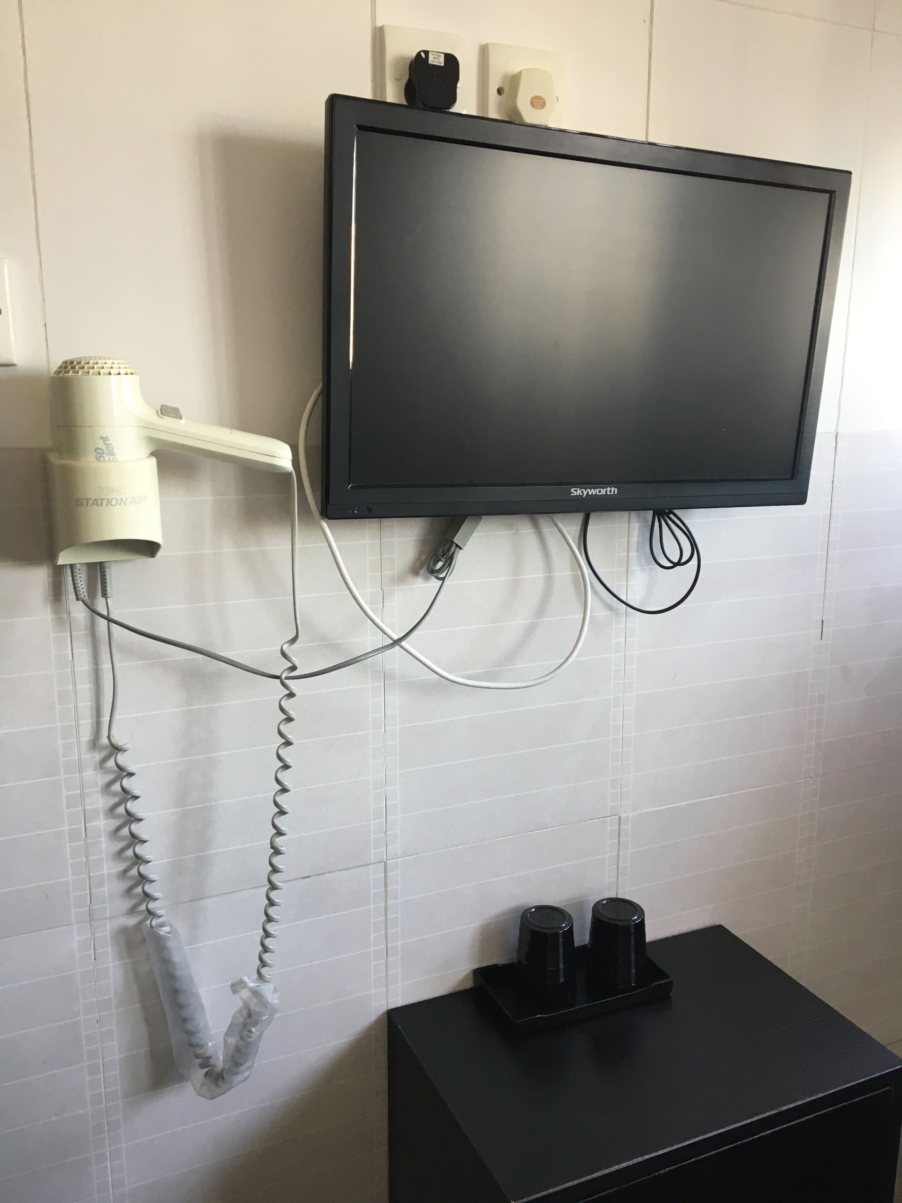 Un secador de pelo, un televisor de pantalla plana y una cafetera sobre una mesa negra en una habitación con paredes de azulejos.