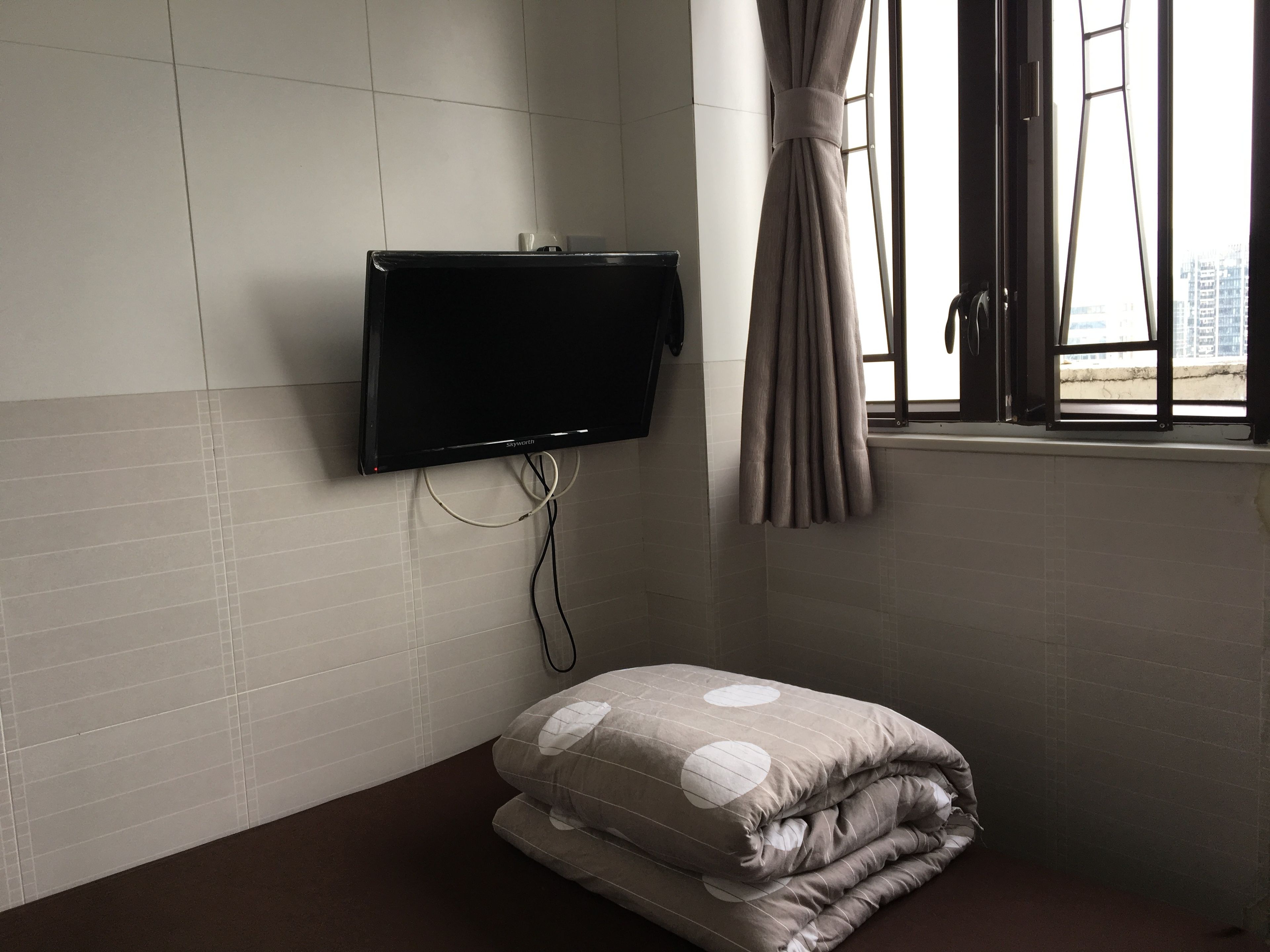 A room with a flat-screen TV mounted on the wall, a window with curtains, and a bed with a polka-dotted blanket.