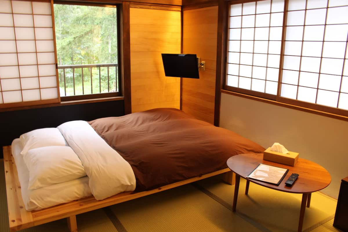 A single bed with a brown blanket and white pillows, a bedside table with a remote control and tissues, and a wall-mounted TV.