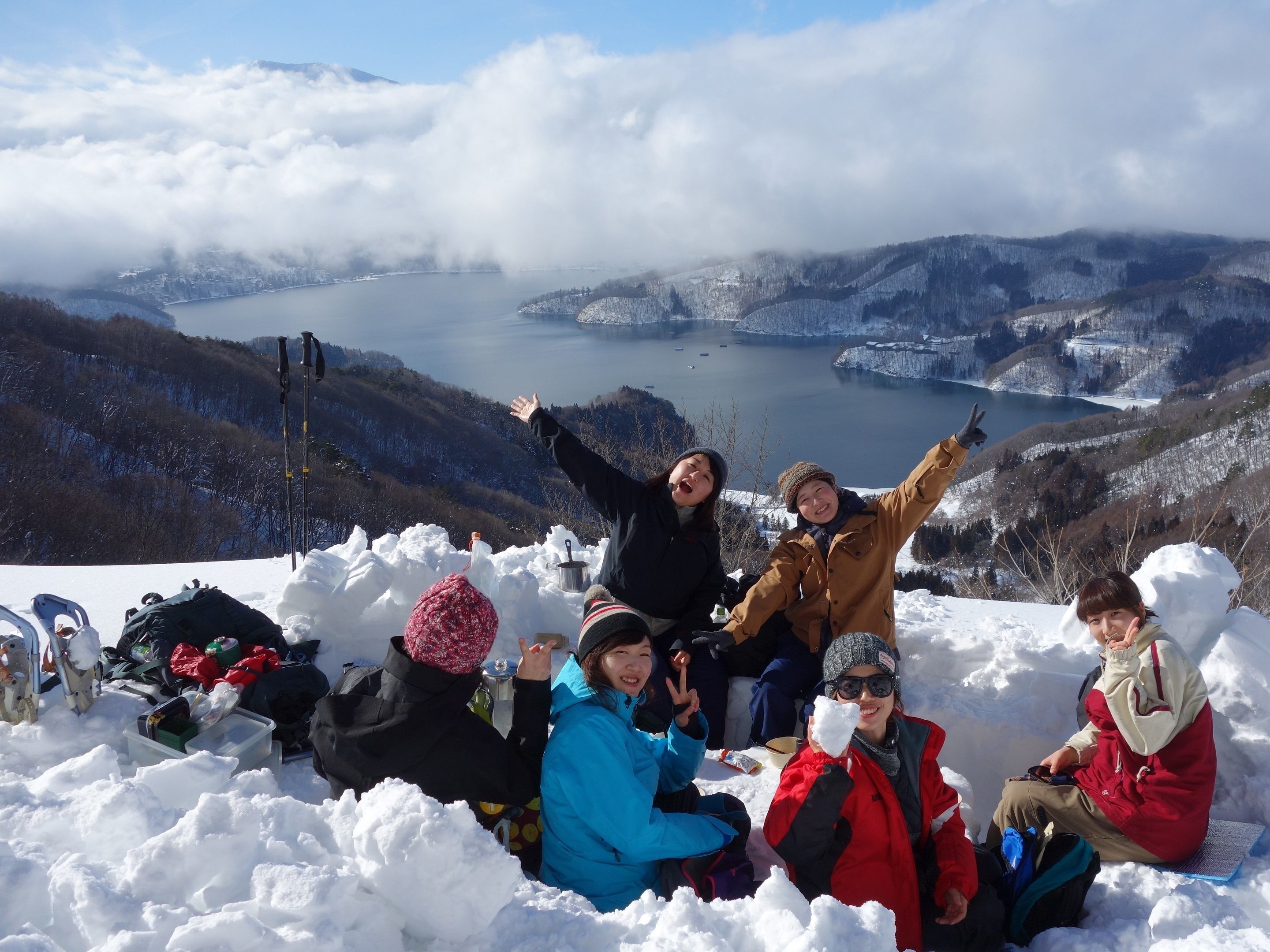 Een groep mensen zit op een besneeuwde heuvel, met een meer en bergen op de achtergrond.