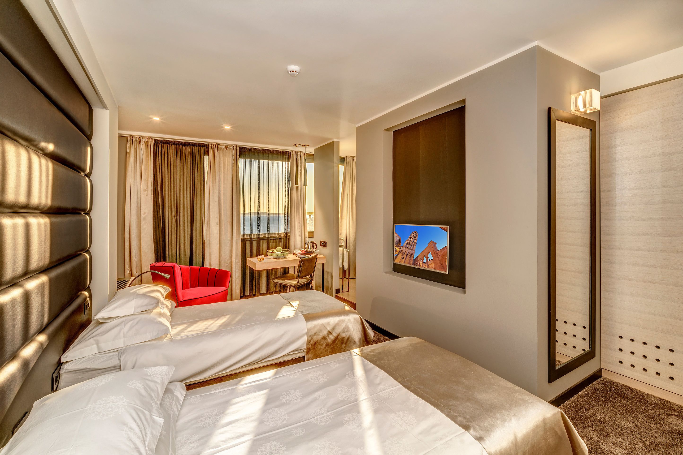 A hotel room with two beds, a desk, a chair, a TV, and a large window with curtains.