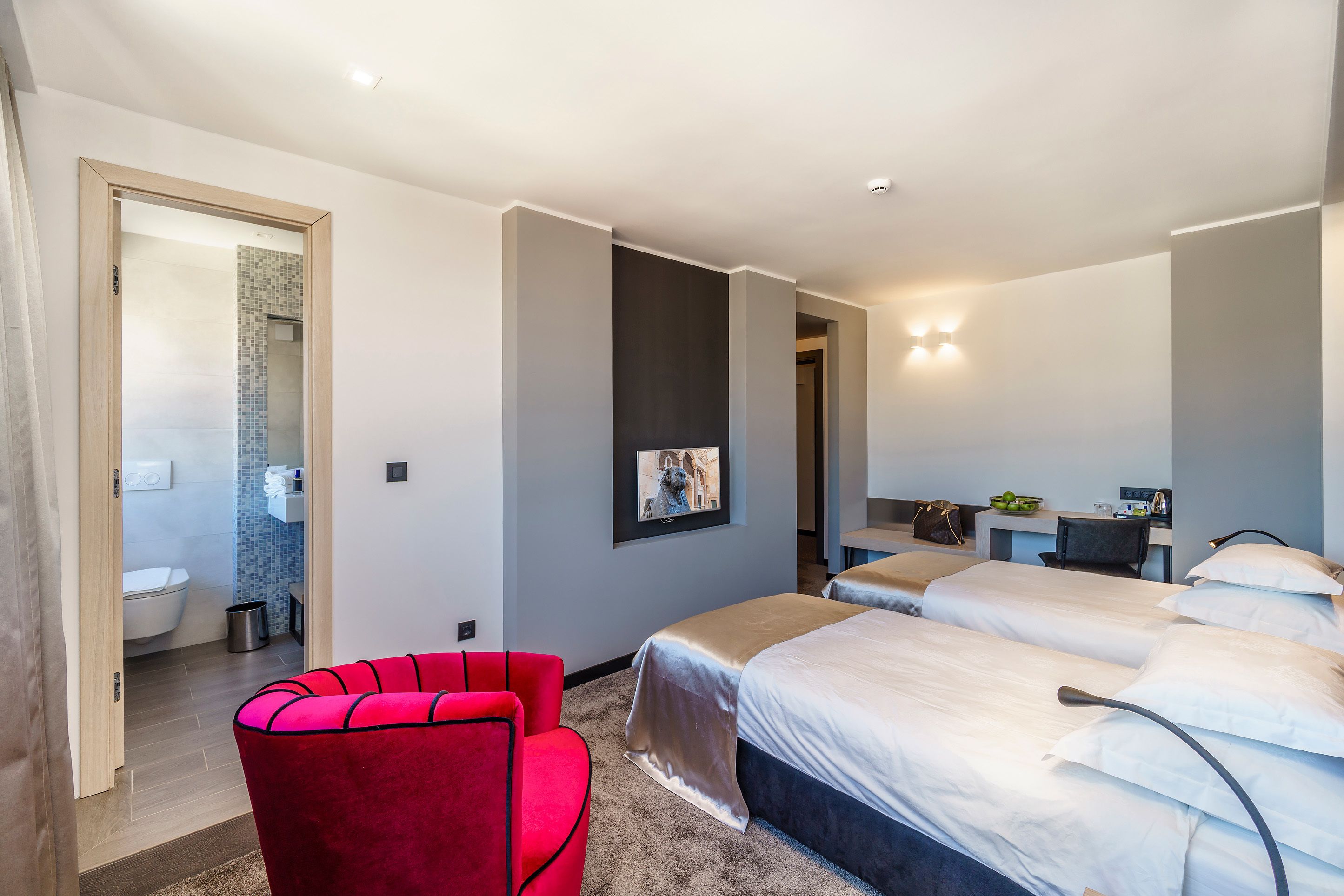 A modern hotel room with two beds, a red chair, a desk, and a bathroom visible through an open door.