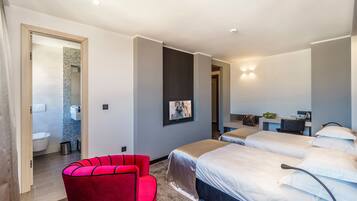 A modern hotel room with two beds, a red chair, a desk, and a bathroom visible through an open door.