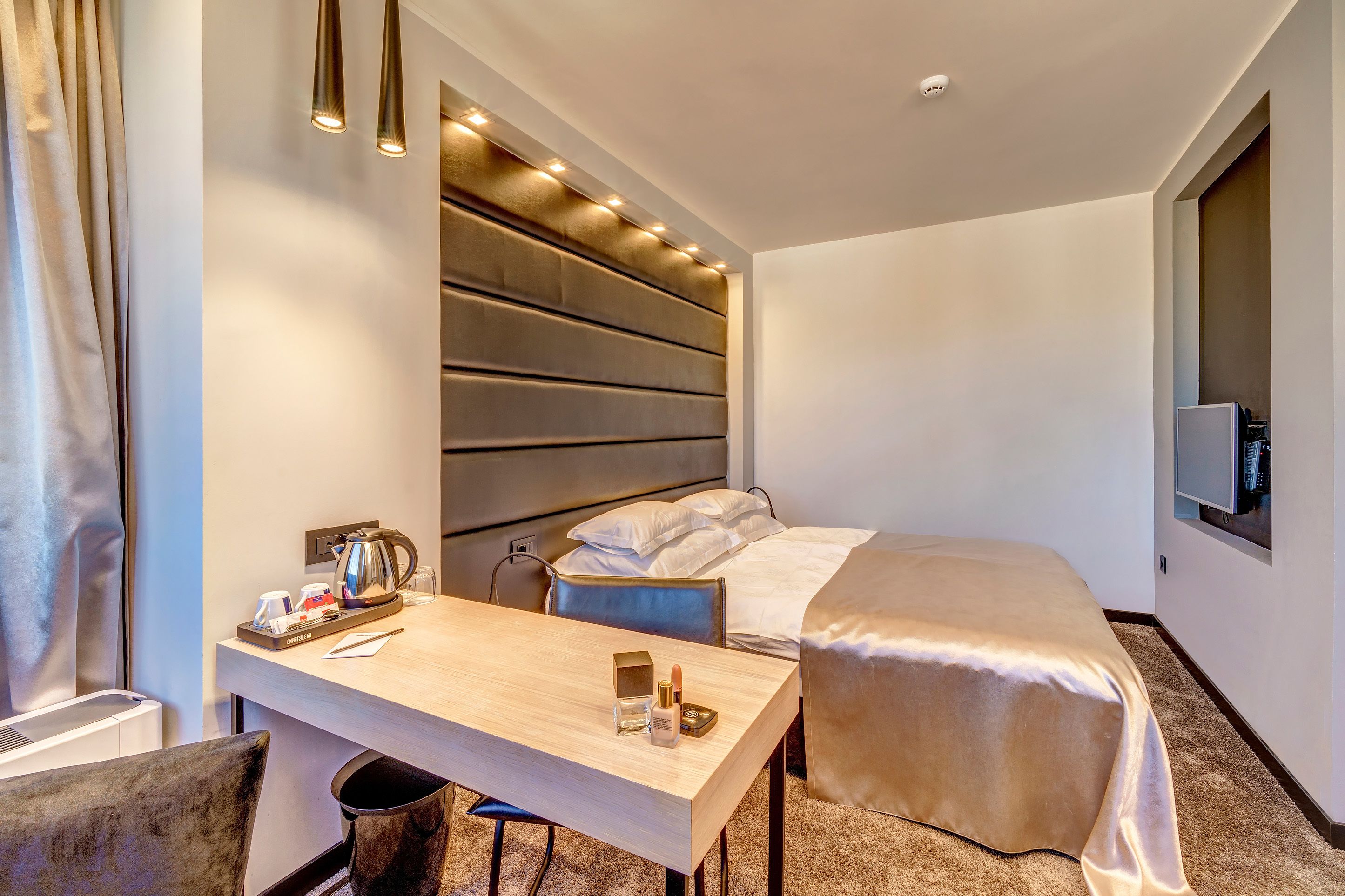 A hotel room with a bed, a desk with a coffee maker, a chair, and a television mounted on the wall.