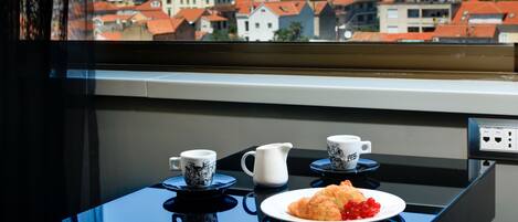 Una mesa con un plato de comida, dos tazas y una jarra, vista a través de una ventana que da a un pueblo con edificios de tejados rojos y montañas al fondo.