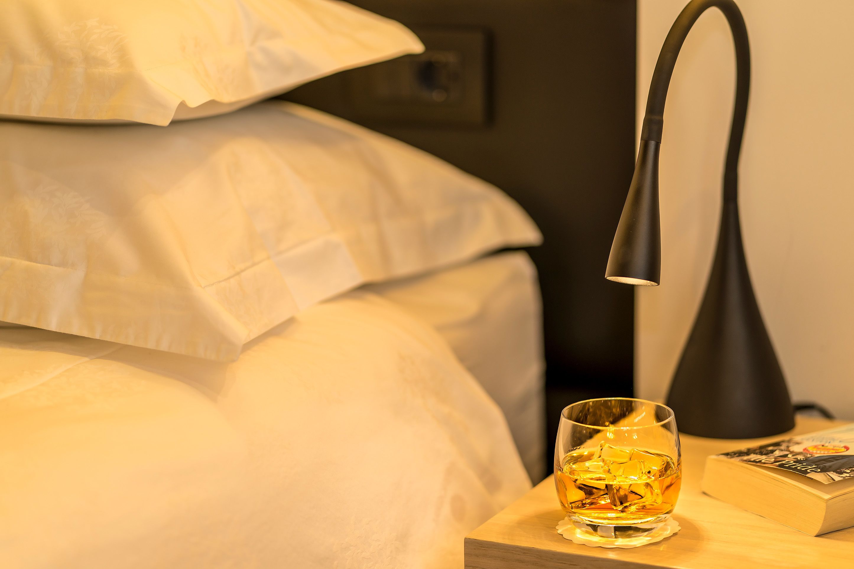 A neatly made bed with white linens, a bedside lamp, a glass of amber liquid, and a book on a wooden nightstand.