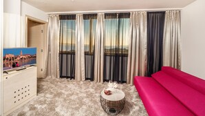 A hotel room with a large window, a sofa, a small round table with a vase, and a television on a cabinet. - The View Luxury Rooms (Split)