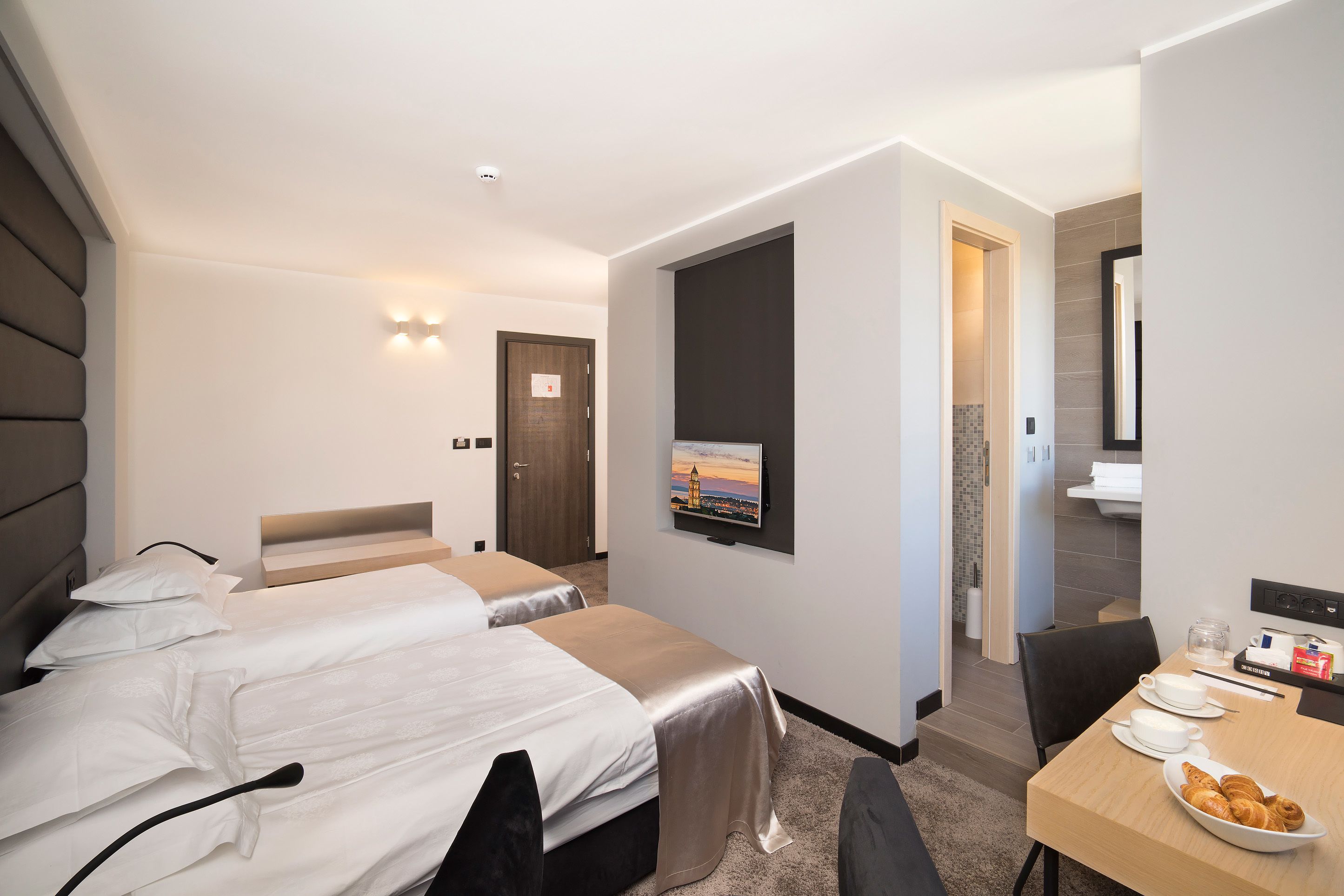 Een hotelkamer met twee bedden, een aan de muur bevestigde televisie, een ontbijttafel met ontbijtartikelen, en een badkamer zichtbaar door een geopende deur.