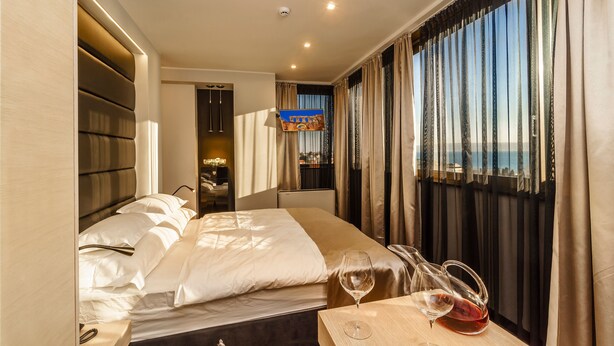 A hotel room with a bed, a bedside table with a wine glass, and a view of the city through large windows. - The View Luxury Rooms (Split)