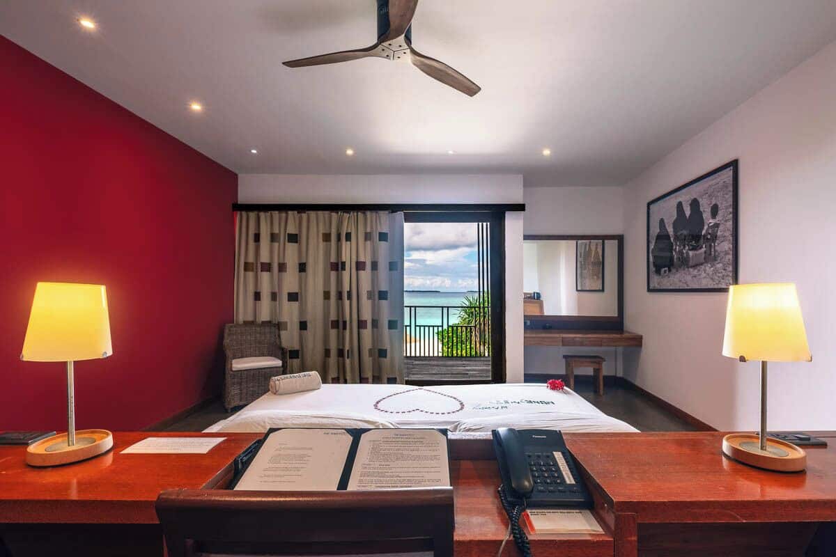 A hotel room with a bed, two bedside lamps, a desk with a telephone, a ceiling fan, a window with curtains, and a view of the sea.